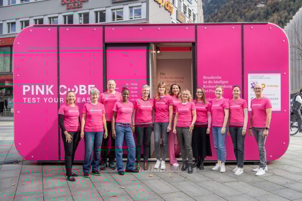 Vor dem leuchtend pinken PINK CUBE Beratungszimmer stehen mehrere Teams in PINK CUBE T‑Shirts bei „Test Your Breast“.
