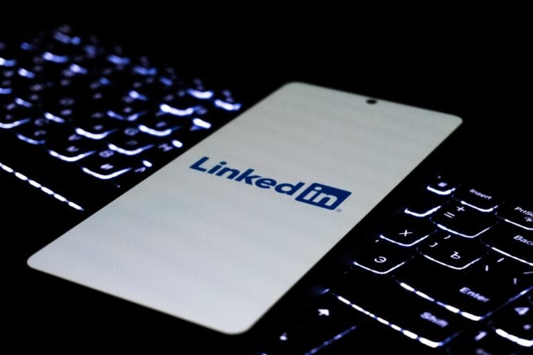 Ein weißes Smartphone mit LinkedIn-Logo liegt auf einer dunkel beleuchteten Laptop-Tastatur, deren Tasten im Halbdunkel blauweiß aufleuchten.