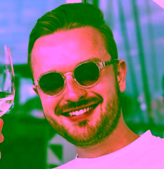 Mann mit blonden Haaren und Sonnenbrille, der ein Glas in der Hand hält und lächelt.