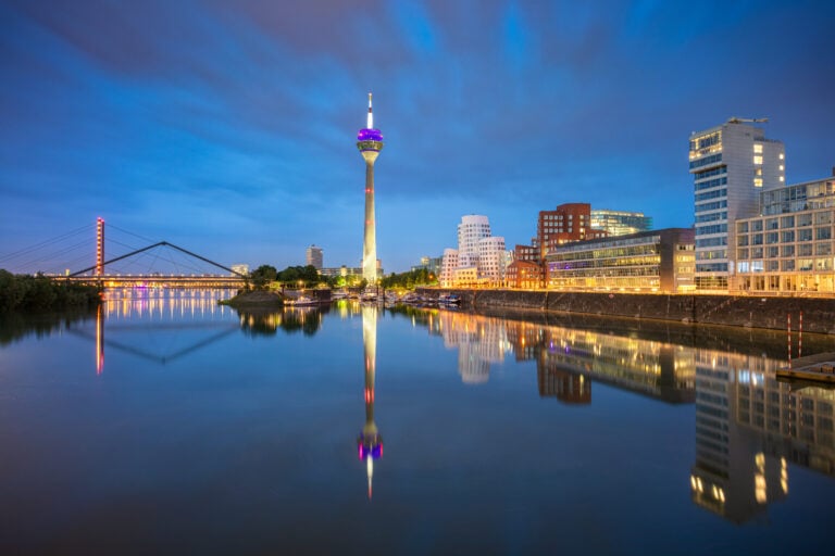 Düsseldorf Skyline mit dem Rheinturm und modernen Gebäuden bei Nacht, reflektiert im Wasser des Rheins.