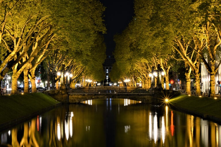 Nachtaufnahme eines beleuchteten Kanals mit Bäumen und Brücke in einer Stadt.