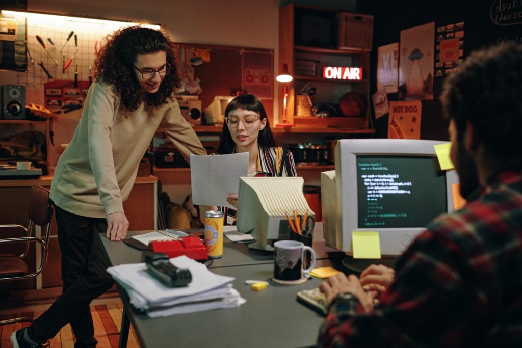 Drei Personen arbeiten in einem unordentlichen Studio/Büro. Im Vordergrund rechts sitzt eine Person vor einem Computer mit Codebildschirm; in der Mitte prüft eine Frau mit Brille Unterlagen, links lehnt eine weitere Person über dem Tisch. Eine rote ON AIR-