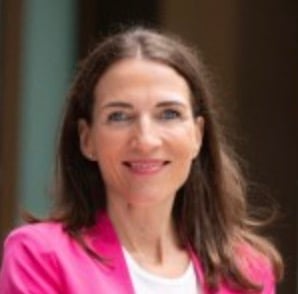 Frau mit langen braunen Haaren und einem rosa Blazer lächelt in die Kamera.